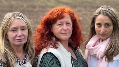 Councillors Mel Allcott, Karen Holmes, and Jessie Jagger. The three councillors are lined up, shoulder to shoulder. Allcott (left) has long blonde hair and is wearing a black shirt with white spots. Holmes has curly ginger hair and is wearing a green and black patterned top with a white gilet over the top. Jagger has dark blonde hair and is wearing a pink scarf and a white top.