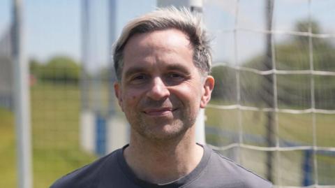 Chris Phillips has dyed blond hair and is wearing a grey T-shirt in front of a football goal.