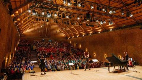 Snape Maltings Concert Hall - a photograph taken from the back of the stage with the auditorium behind. A man at a grand piano is on the right, with three people, standing apart at the front of the stage, holding their arms aloft. There is a full house.