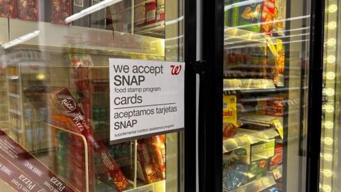 A sign on a grocery store fridge reads "We accepts SNAP food stamp program' in both English and Spanish. Frozen food is seen in the background. 