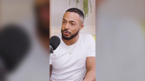Tyler West sits in a white t-shirt with a black mic in front of him. A hanging plant is behind him.