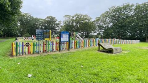 A multicoloured fence encloses a play area. In the distance you can see a slide and a climbing frame. It is in a grassy area.