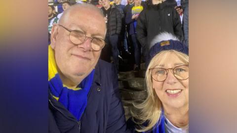 Ged has short grey hair and is wearing dark rimmed glasses and a blue jacket with a blue and yellow Warrington Wolves scarf. Jean has long blonde hair and is wearing dark rimmed glasses and a wooly blue hat with a white bobble. They are stood at a stand full of Warrington Wolves fans.