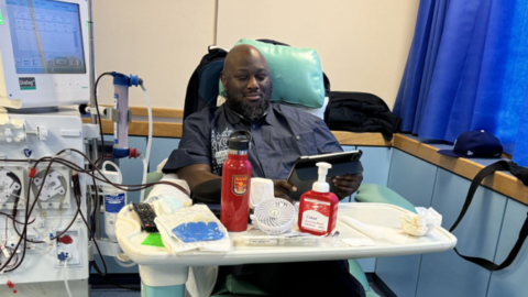Edmund seated in a hospital treatment chair next to a dialysis machine, with medical tubing, a tray of supplies, and a tablet resting on their lap in a clinical setting.