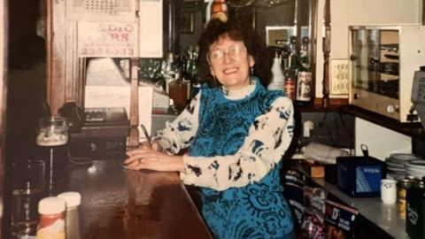 Nancy Swanick, with a blue dress with white sleeves, smiles behind the bar in the 1980s
