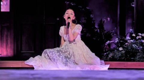 Ariana Grande sits on the stage, surrounded by flowers, as she sings on her Eternal Sunshine tour