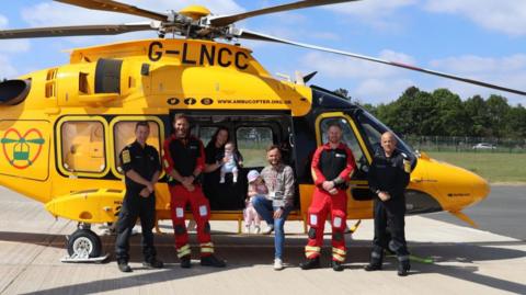 Four men from an air ambulance crew pose with a family in front of a yellow helicopter on a runway at an airfield. Two men wear red and black paramedic uniforms, while two others wear black flying overalls. The family, including a man, woman, young girl and baby boy, are casually dressed and sitting within the open door space of the helicopter.