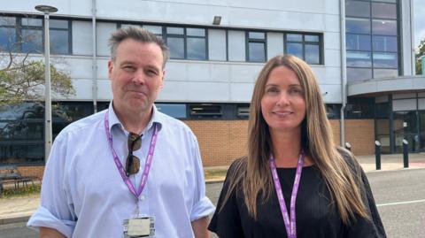 Wood and Davies photographed side-by-side. Wood is wearing a blue collared shirt, and Davies is wearing a black dress. They are both wearing purple lanyards. They are stood outside one of the college buildings, a large multi-storey structure with big glass windows. 
