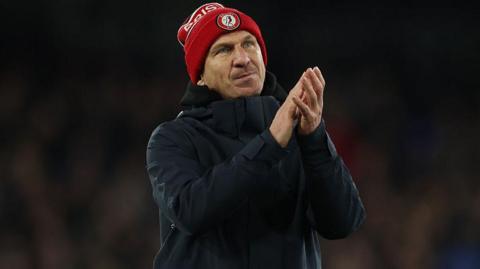 Gerhard Struber wearing a red Bristol City beanie hat and dark overcoat applauds supporters