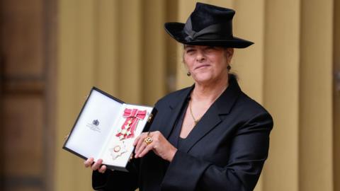 Dame Tracey Emin wearing a black suit and big hat holding her damehood medal 