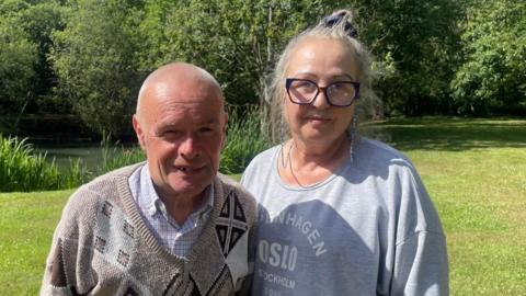 A man wearing a grey jumper with patterns and a woman wearing a grey top stand together outside. Grass and trees can be seen behind them.