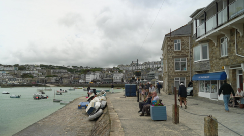 A harbour scene shows a stone promenade lined with buildings, with small boats floating on calm water and a town spread across the hillside in the background under a cloudy sky. People are sitting and walking along the waterfront near stacked dinghies, while a gallery and other seaside shops face the quay.