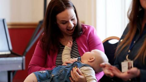 Kate Forbes, in pink blazer and black and white stripe top, sits in a mother and baby group, holding her baby daughter in her lap and smiling down at her.