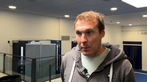 A middle-aged man with short hair wearing a hoodie and t-shirt in a school building.