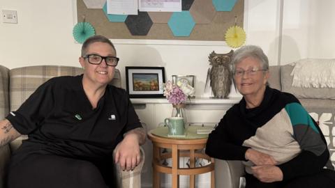 A woman with black glasses and short hair and an elderly woman with glasses and short grey hair. They are sat in arm chairs and smiling at the camera in front of them