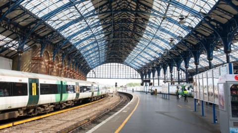 A train station. The roof is made of blue steel bars. A white and green train is visible. 