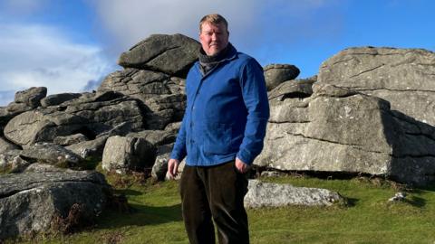 Artist and author, Alex Murdin, is wearing a blue jacket, and stands in front of Bonehill Rocks on Dartmoor. The grass is bright green and the sky is blue with some light clouds. 