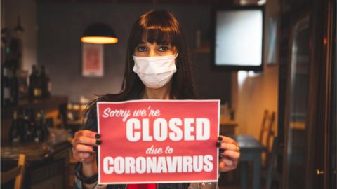 Woman in bar holding sign saying "Sorry we're closed due to coronavirus"