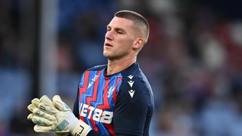 Sam Johnstone with hands together, in goalkeeping gloves, while he looks off camera. He is wearing a Palace training top which has blue sleeves and a red and blue zigzag pattern on the front