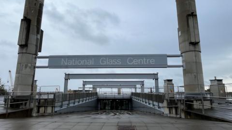 The entrance of the National Glass Centre in Sunderland. A large grey sign is suspended between two high cement pillars. A sloped path leads to the glass doors of the building. The fenced off roof of the building is visible in the distance.
