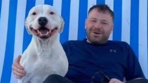 Paul Scott sits against a blue and white striped background, with a white bulldog at his side. He is smiling and is wearing a blue top and black trousers 