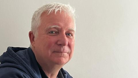 A man with white hair. He is wearing a hooded top.