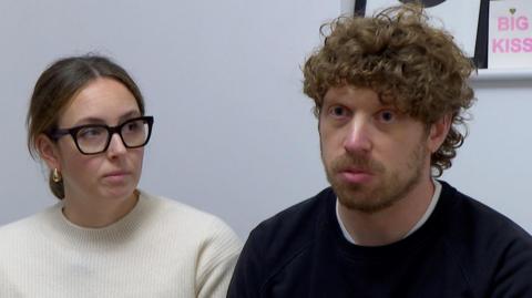 Luke and Lucy sitting together. Luke is wearing a black jumper and Lucy is wearing a white jumper.  