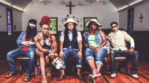 A promotional photo of Arrested Development. Three men and two women sit on a bench inside a small chapel, which has crosses on the wall and stained glass windows. They all pose and look at the camera with serious expressions. They wear denim jeans and two have sunhats on. 