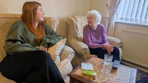 Two women are seated and engaged in conversation. On the left, a younger woman with long, red hair wears a green jumper and black trousers. Opposite her, an older woman with short white hair wears a purple sweater and black trousers. In front of them is a coffee table with a glass of water and remote control on. 
