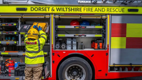 A firefighter stands facing a fire engine. They are wearing a hi-vis jacket and a yellow helmet. They are loading kit onto the fire engine. At the top of the fire engine are the words Dorset and Wiltshire Fire and Rescue.