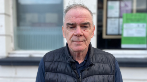 Noel Kelly pictured with a neutral expression on his face. He is wearing a black body warmer on top of a long sleeved blue shirt.