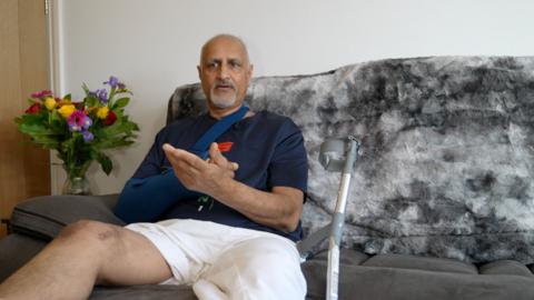 A man with short grey hair and a beard sitting on a dark grey sofa with a dark blue shirt and one bare leg up on a stool with a silver crutch beside him