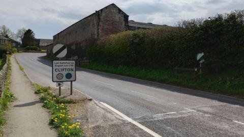 The A6 road leading into the village of Clifton of Penrith. The national speed limit signs have been turned around so the signs lead into the village instead of out. The signs read CLIFTON and there are bushes and dandelions on either side of the road.