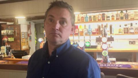 A man wearing a blue shirt is standing in front of a brightly-lit bar