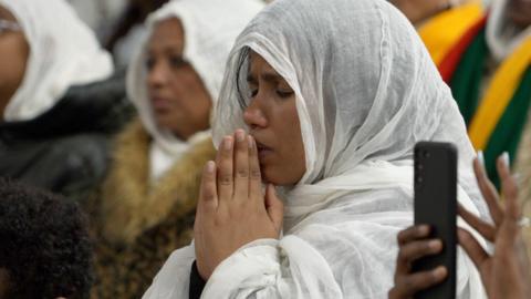 A woman with a white shawl over her head and body has her hands in front of her mouth in a praying position. She has her eyes closed.