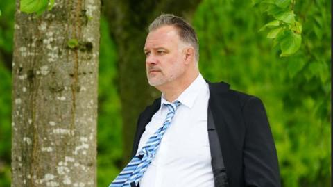 A man with receding black and grey hair and grey beard, wearing a white shirt, blue tie and black blazer. He is stood next to to a tree looking away