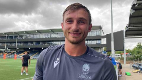Pictured is Josh Bassett. He is smiling and has a blonde stubbly beard and wet dark blonde hair. He is wearing a grey sport t-shirt with his initials, JB, printed on the left hand side of his shirt in a light grey colour. He is stood on a rugby pitch where two people are talking in the background.