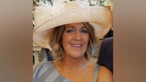 Jane Blackwell smiling at the camera. She has curly, shoulder-length blonde hair, and is wearing a large cream hat with feathers and a grey formal dress.