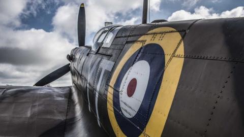 A close-up view of a the World War Two aeroplane the Spitfire, showing the left side painted grey. There is a yellow, blue, white and maroon circular log on its side above its wing. Beyond is the clear glass/plastic of the cockpits and its propeller. 