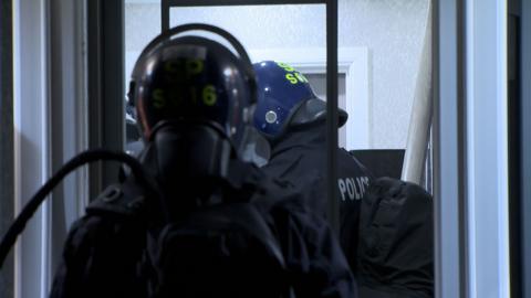 Police officers wearing black tactical clothing and helmets with yellow writing, carry riot shields as they enter a house
