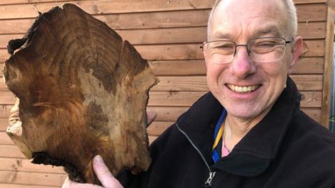 Dave Everett who has short white hair, rectangular glasses and is wearing a dark blue fleece. He is holding up a slice of tree and is smiling broadly