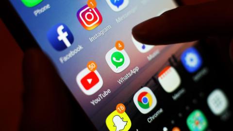 Close-up of a smartphone screen displaying social media and messaging apps, including Instagram (with one notification), Facebook, WhatsApp (four notifications), YouTube (50 notifications), Snapchat (16 notifications) and Messenger. A finger hovers over the screen, appearing about to tap an app.