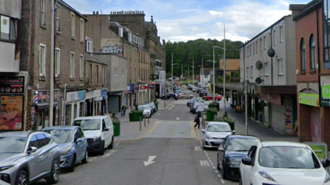A high street with shops on each side. Cars are parked on either side of the road.