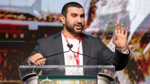 Hamdi speaking at a podium with with hand up to illustrate a point