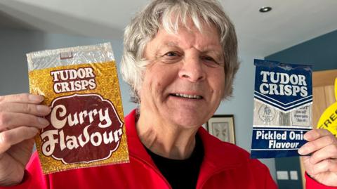 Frank Munford is holding an empty packet of vintage Tudor Crisps in each hand. On the left is a curry-flavoured packet and on the right is a pickled onion-flavour bag. He is holding them proudly, smiling at the camera. He is  wearing a vintage red and white Walkers Crisps tracksuit top.