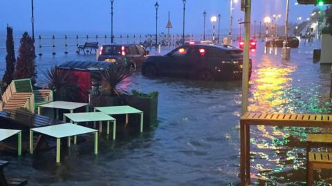 A photograph of the flooded Penarth sea front. Four cars are trapped in the water.