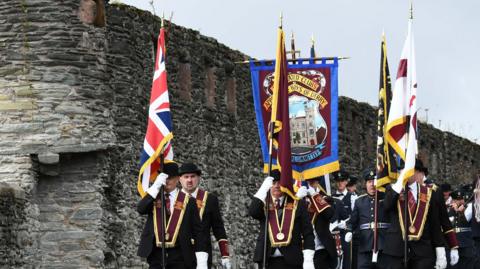 A group of men wearing black suits, black bowler hats, maroon sashes and white gloves parade next to the City of Derry's walls. There are a number of flags