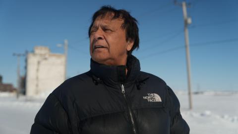 An image of Mike Pence, mayor of Churchill. He is wearing a black 'The North Face' puffer jacket. Behind him is the Port of Churchill blanketed with snow, and a clear blue sky above. He is looking towards the left.
