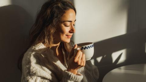 A woman drinks a coffee sat in a kitchen with morning sun light shining in through a window