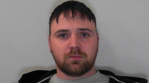 Ashley Watson is looking at the camera. He has a short dark beard and short dark hair and is wearing a black hooded jacked over a grey t-shirt
